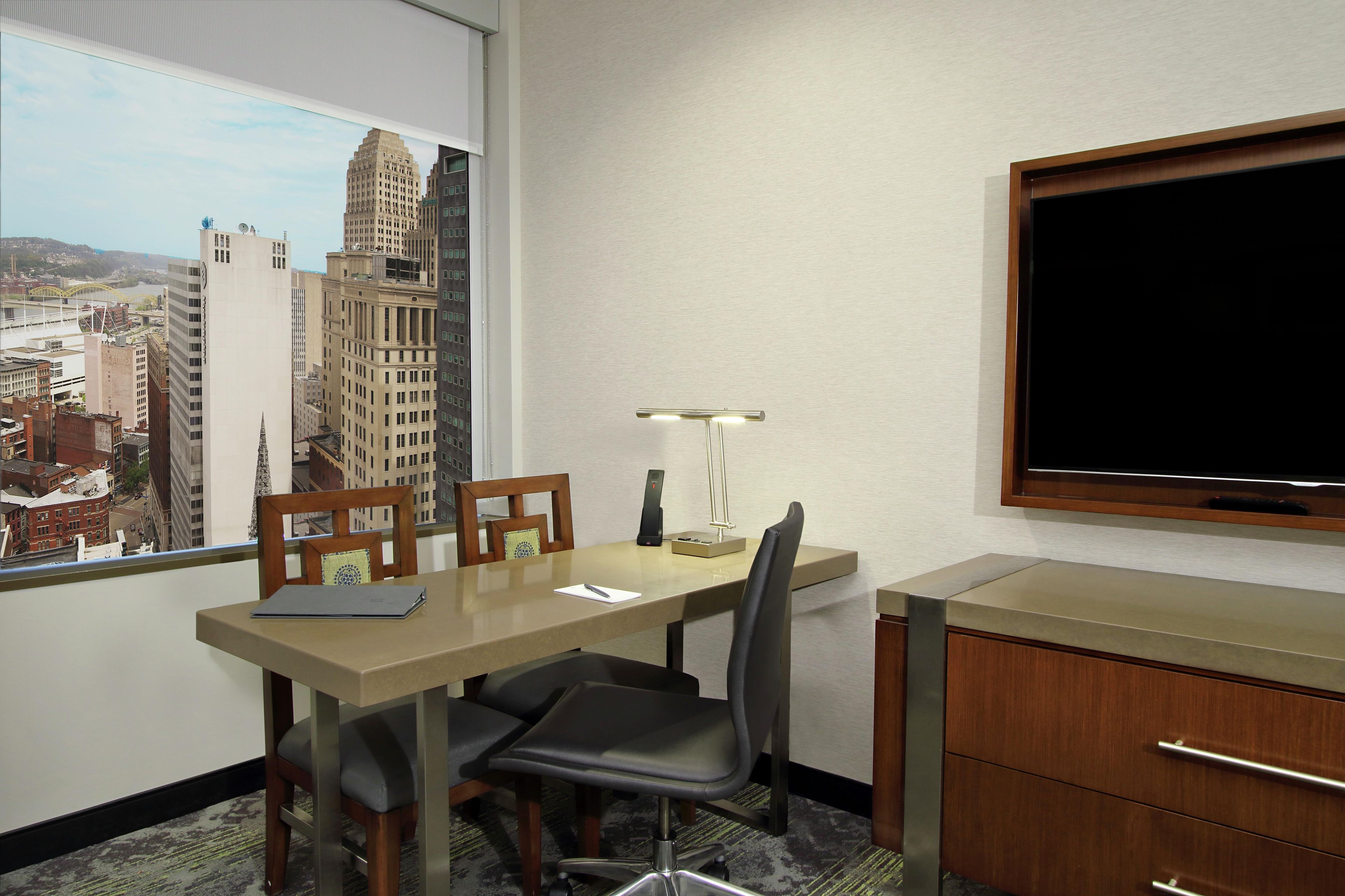 A hotel room with a desk, two chairs, a lamp, a TV, and a view of a cityscape.