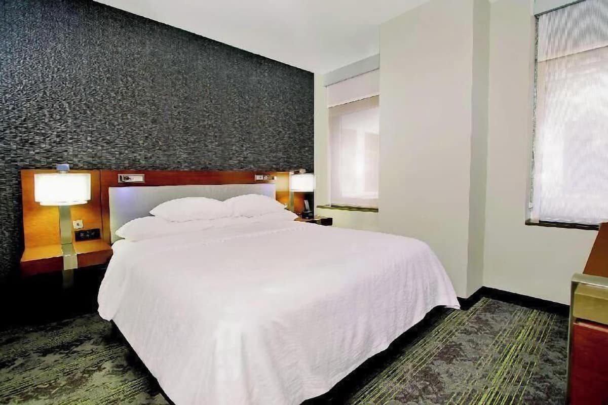 Habitación de hotel con una cama grande, dos lámparas de noche, un cabecero, una ventana con cortinas y piso alfombrado.