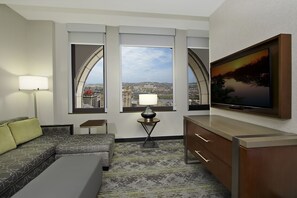 A hotel room with a sofa, a small table with a lamp, a TV on a stand, and a large window offering a city view. - Embassy Suites By Hilton Pittsburgh Downtown (Pittsburgh)