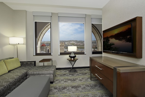 Ein Hotelzimmer mit einer Couch, einem kleinen Tisch mit Lampe, einem Fernseher auf einem Schrank und einem großen Fenster mit Blick auf die Stadt.