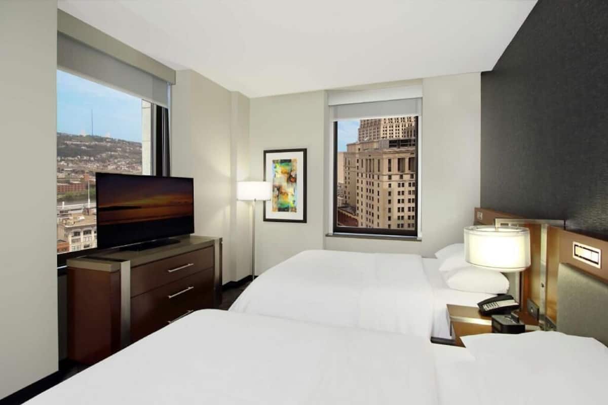 Habitación de hotel con cama, televisor, escritorio con cajones, lámpara, teléfono y ventana con vista a la ciudad.