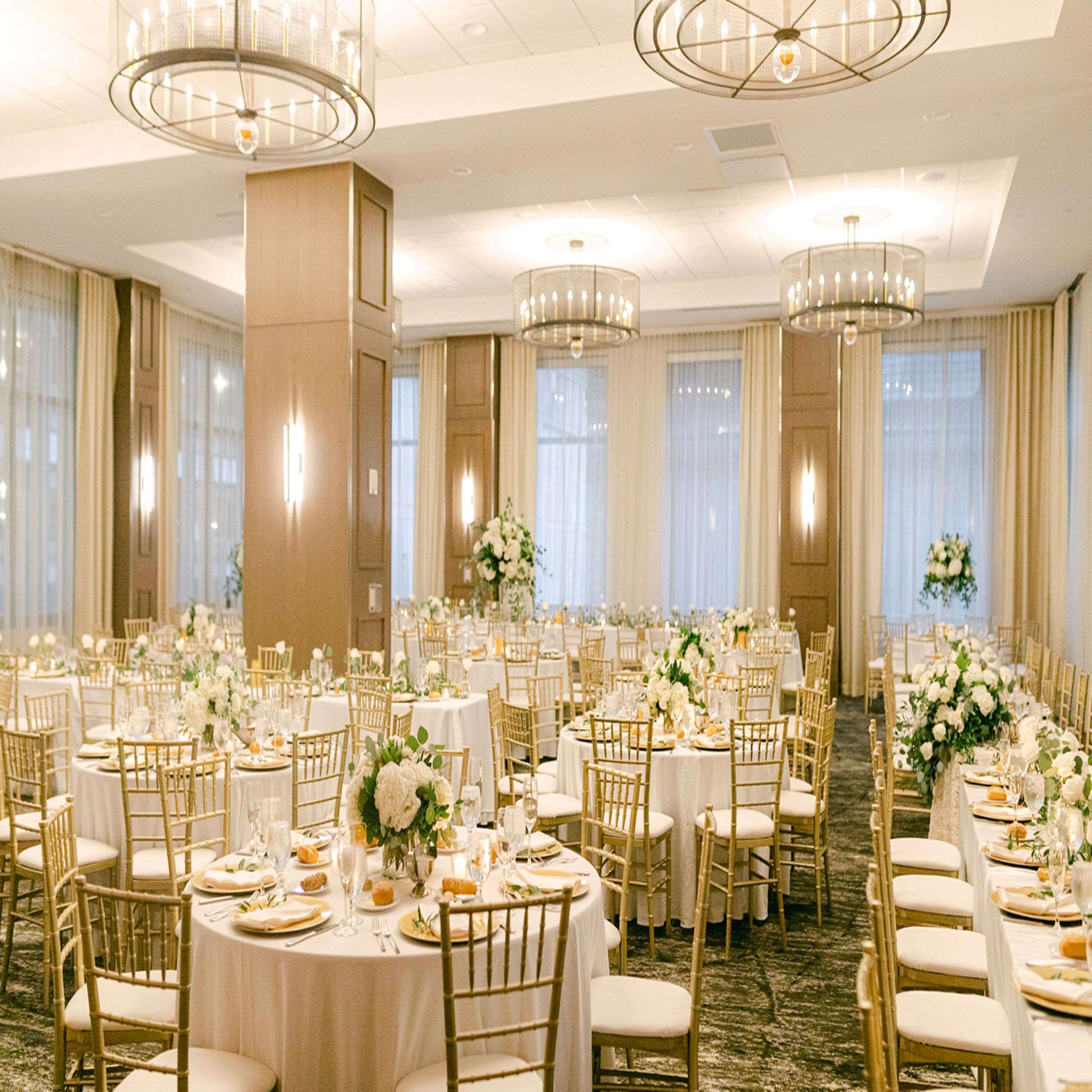 A banquet hall with round tables, gold chairs, and chandeliers.