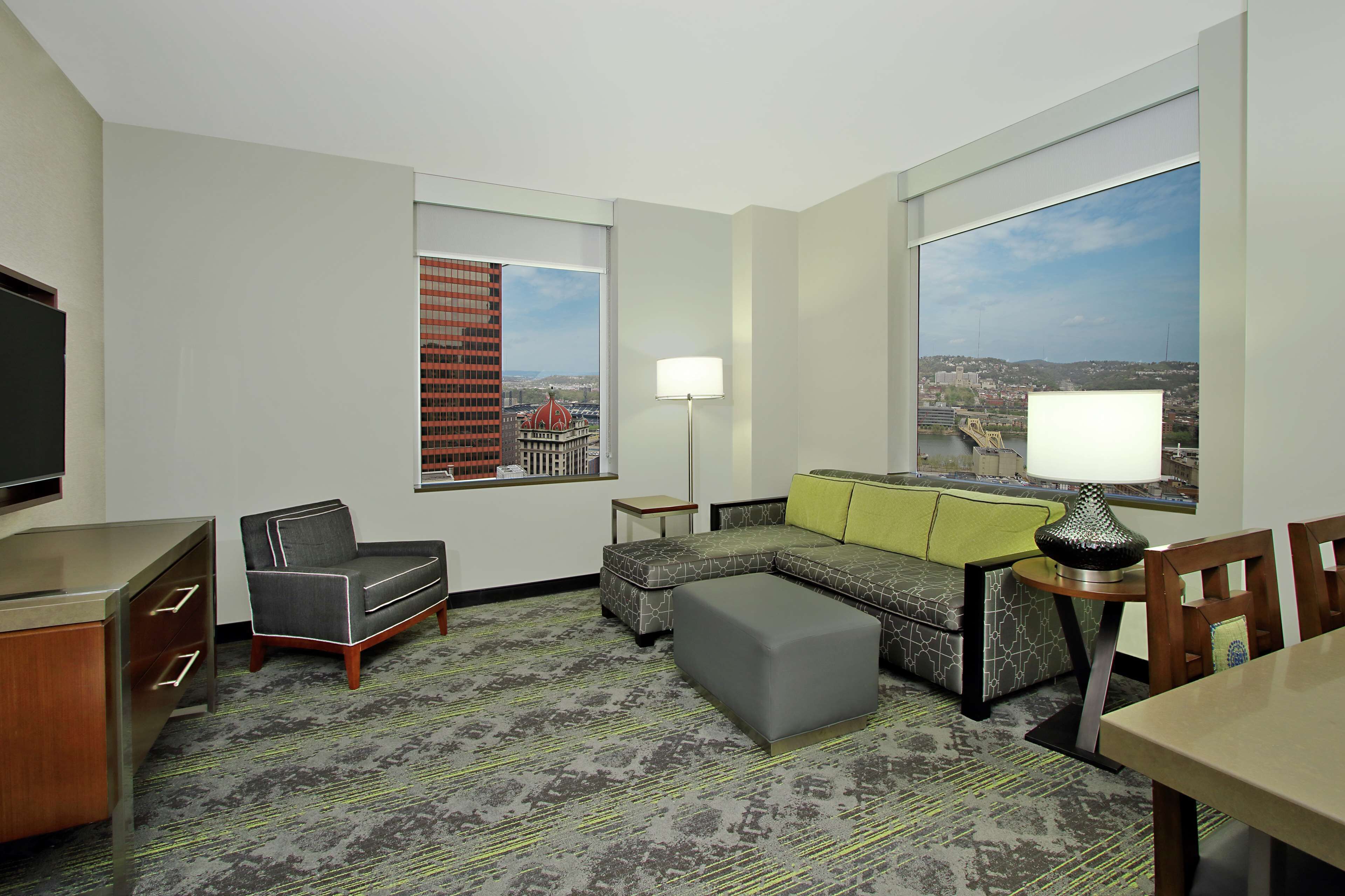 A hotel room with a sofa, ottoman, TV, lamp, and a city view.