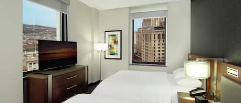 A hotel room with a bed, a TV on a dresser, a painting, and a window with a city view.