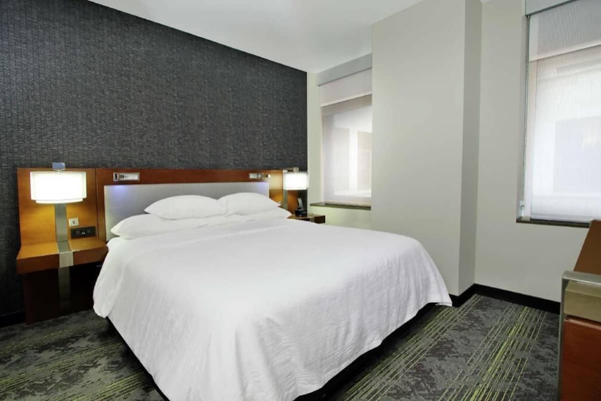 Habitación de hotel con una cama grande, dos mesitas de noche con lámparas, un cabecero, una ventana con persianas y suelo alfombrado.