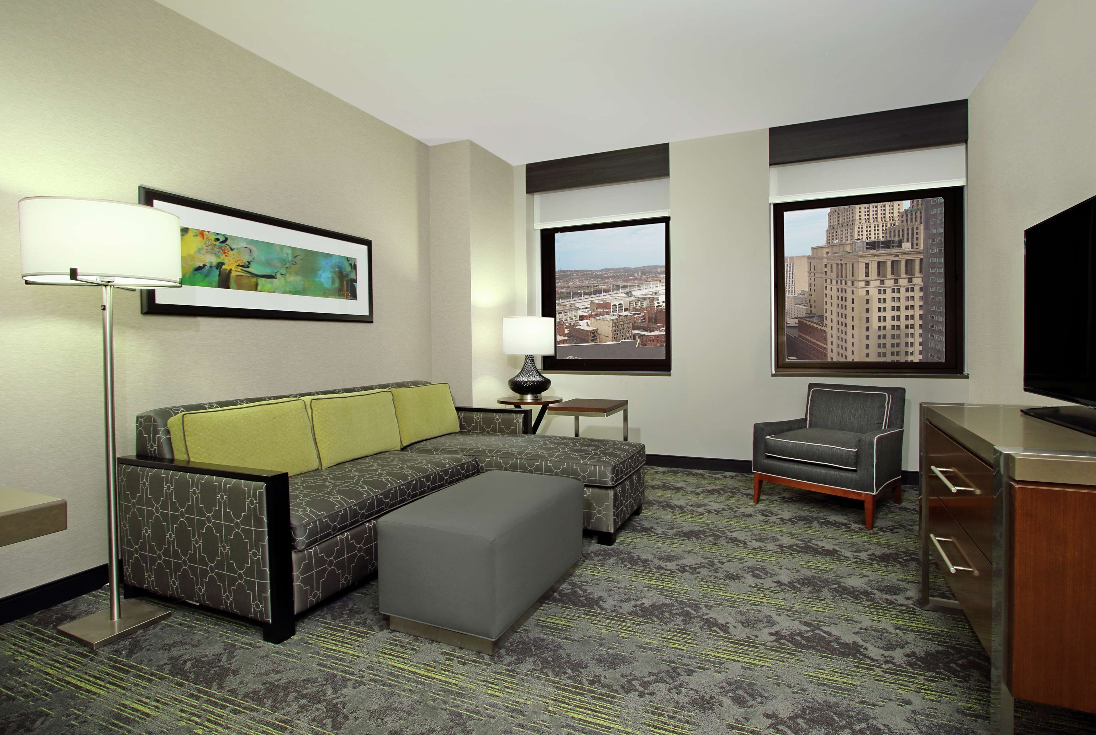A hotel room with a sectional sofa, a single chair, a desk with a TV, a lamp, and a painting on the wall.