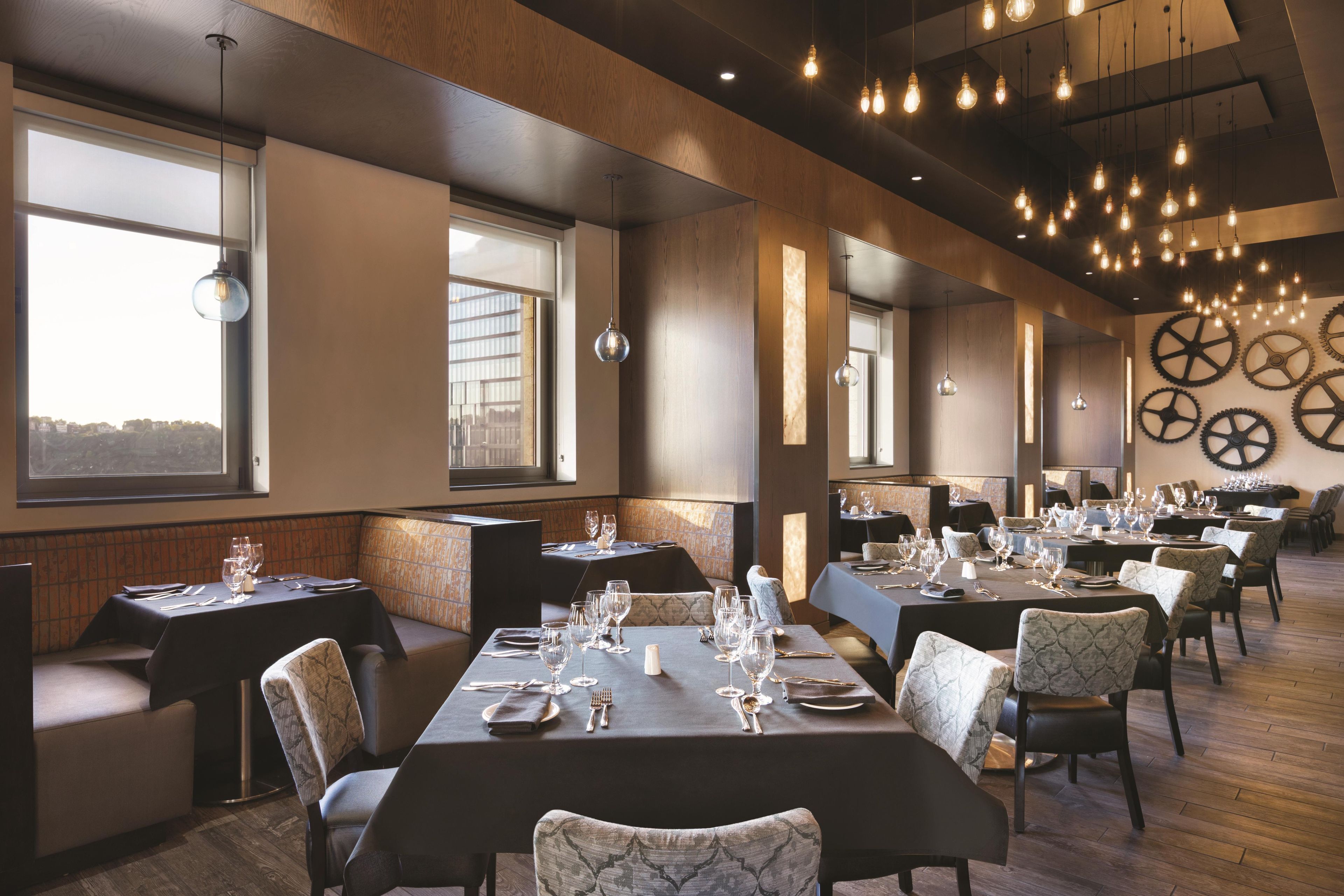 A restaurant with multiple dining tables set for guests, large windows, and a ceiling with hanging light fixtures.