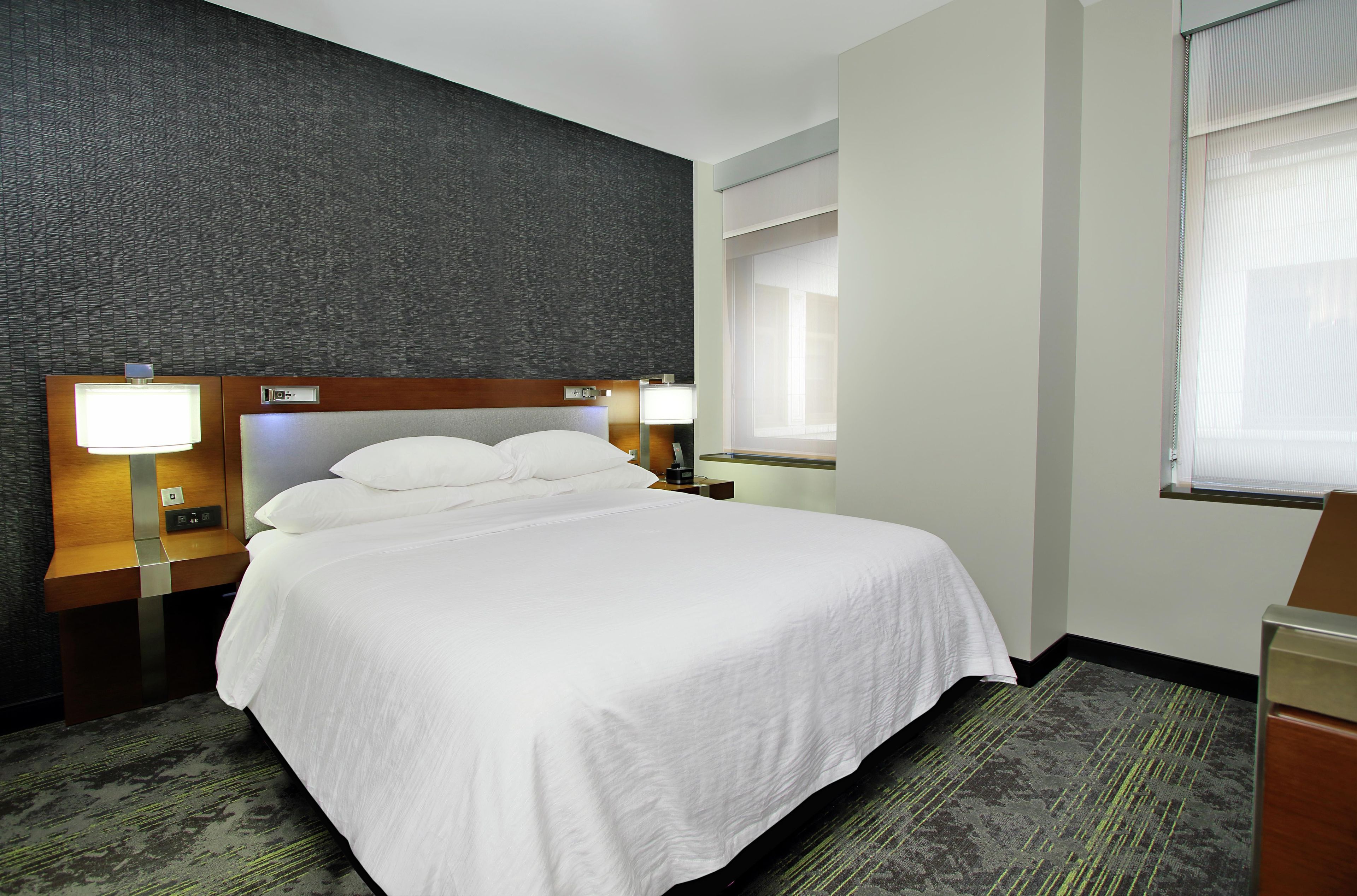 A hotel room with a large bed, two bedside tables with lamps, a window with blinds, and a textured wall.