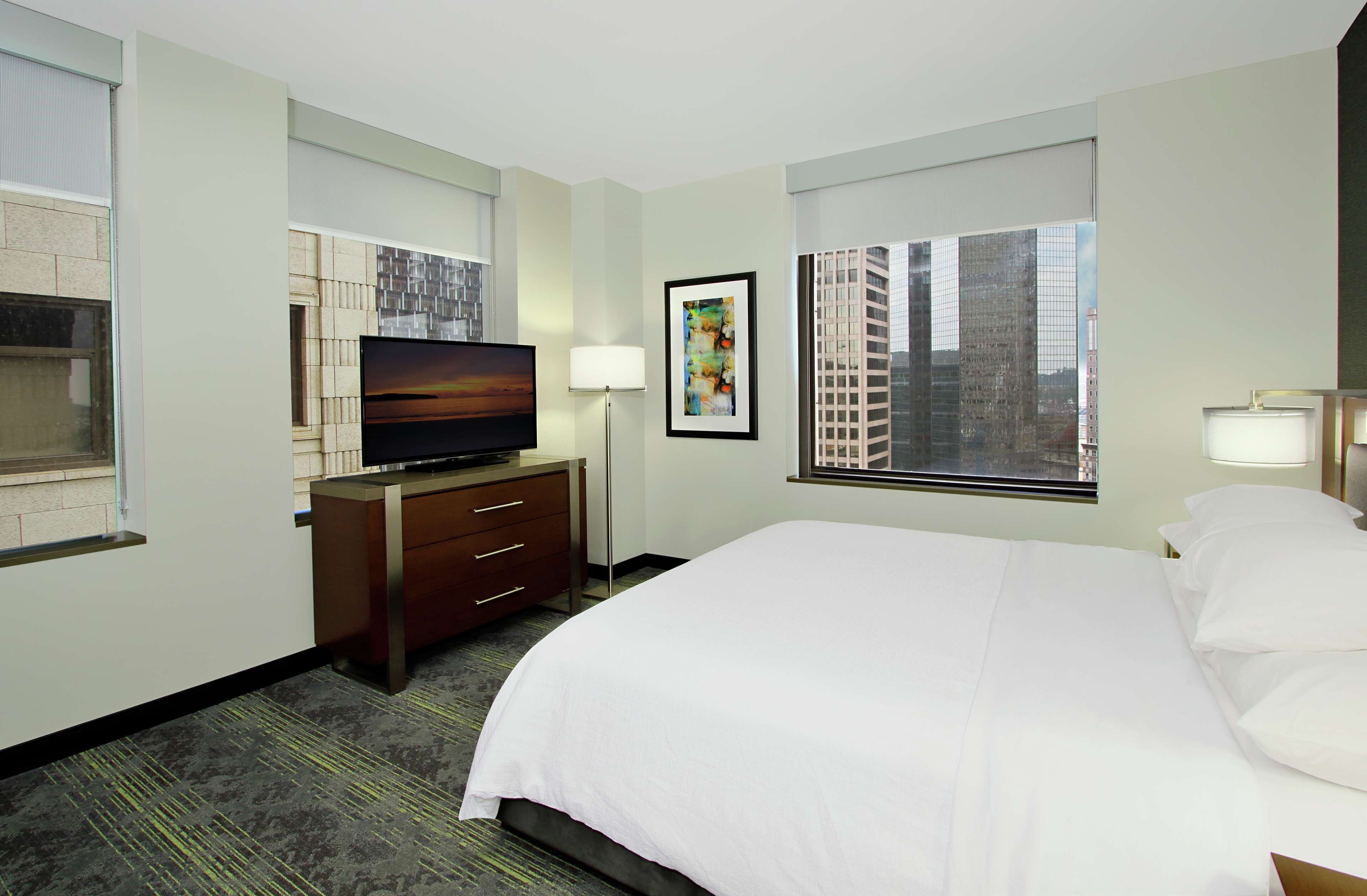 Habitación de hotel con una cama grande, un televisor de pantalla plana, un escritorio con cajones, una lámpara y una ventana con vista a la ciudad.