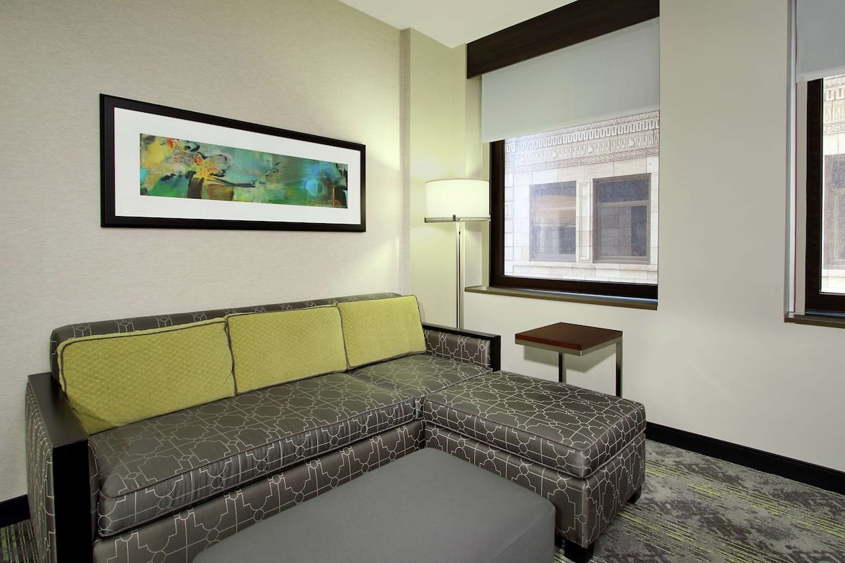 Una habitación de hotel moderna con un sofá seccional, una mesita auxiliar, una lámpara de pie y un cuadro abstracto enmarcado en la pared.