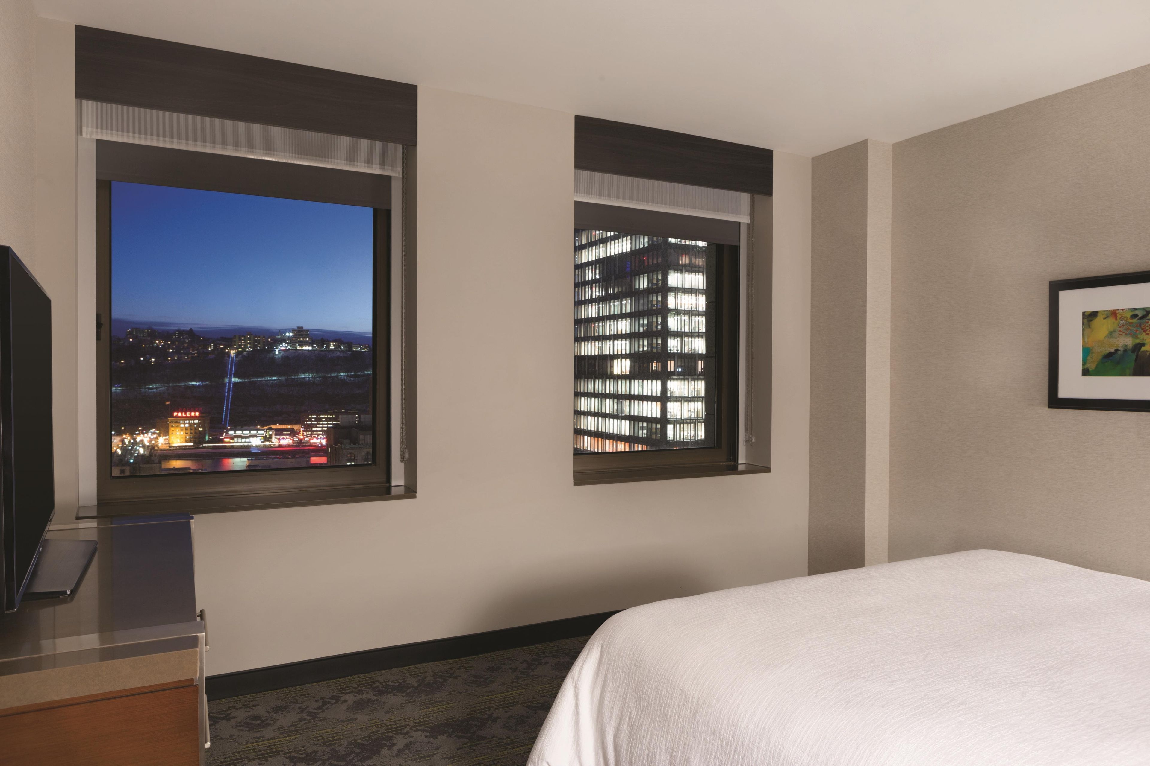A hotel room with a bed, a desk, and two windows offering city views.