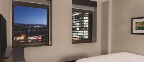 A hotel room with a bed, a desk, and two windows offering city views.