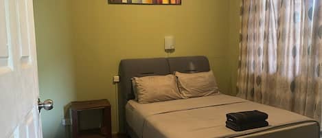 A bedroom with a bed, a nightstand, a wall-mounted light, and a window with curtains.
