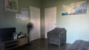 A room with a television, a fan, a grey armchair, and a painting on the wall. - Ceaser's Place (Ranau)