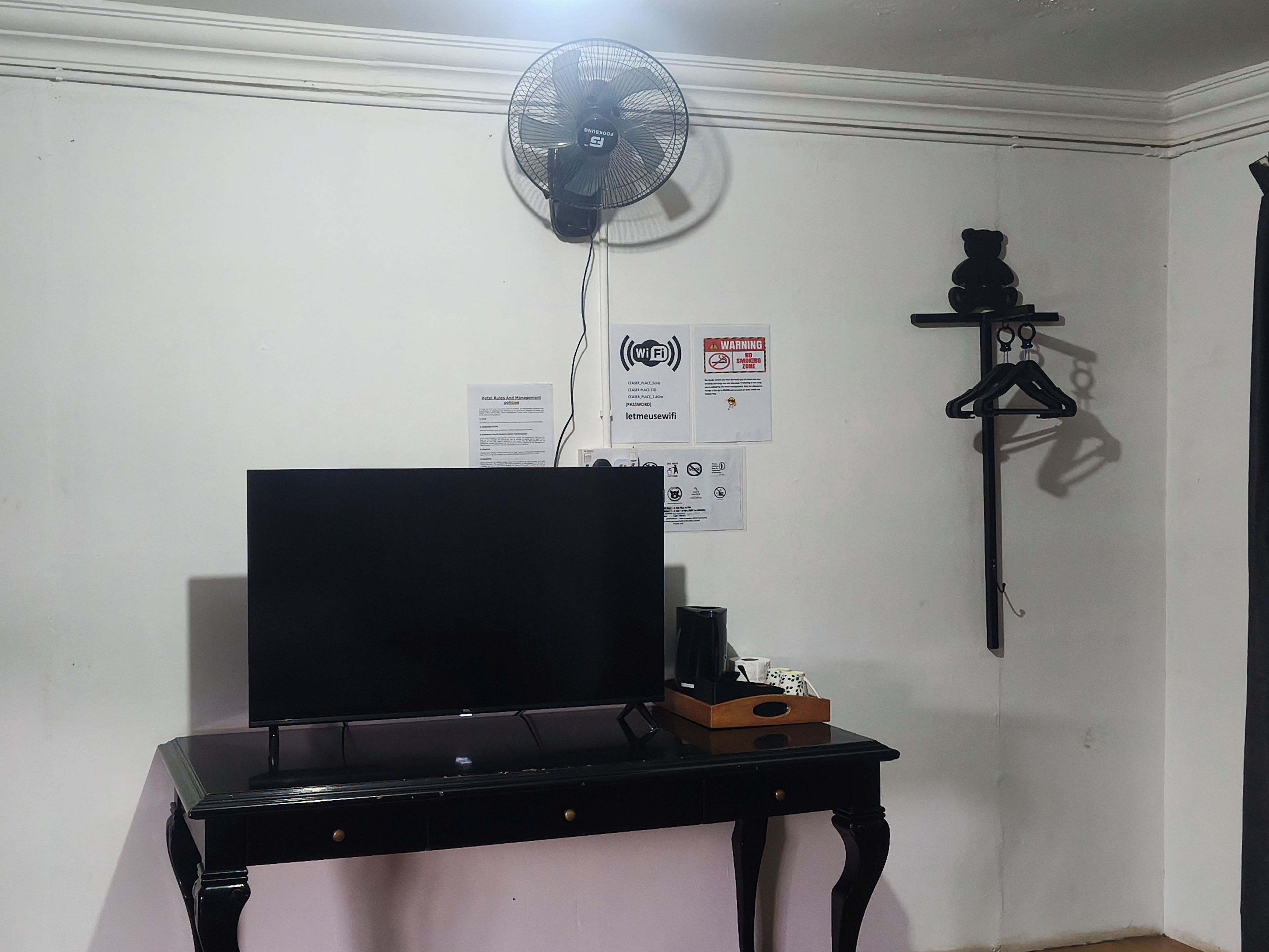 A room with a black desk, a wall-mounted fan, and a shelf with a hanging item.