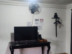 A room with a black desk, a wall-mounted fan, and a shelf with a hanging item. - Ceaser's Place (Ranau)