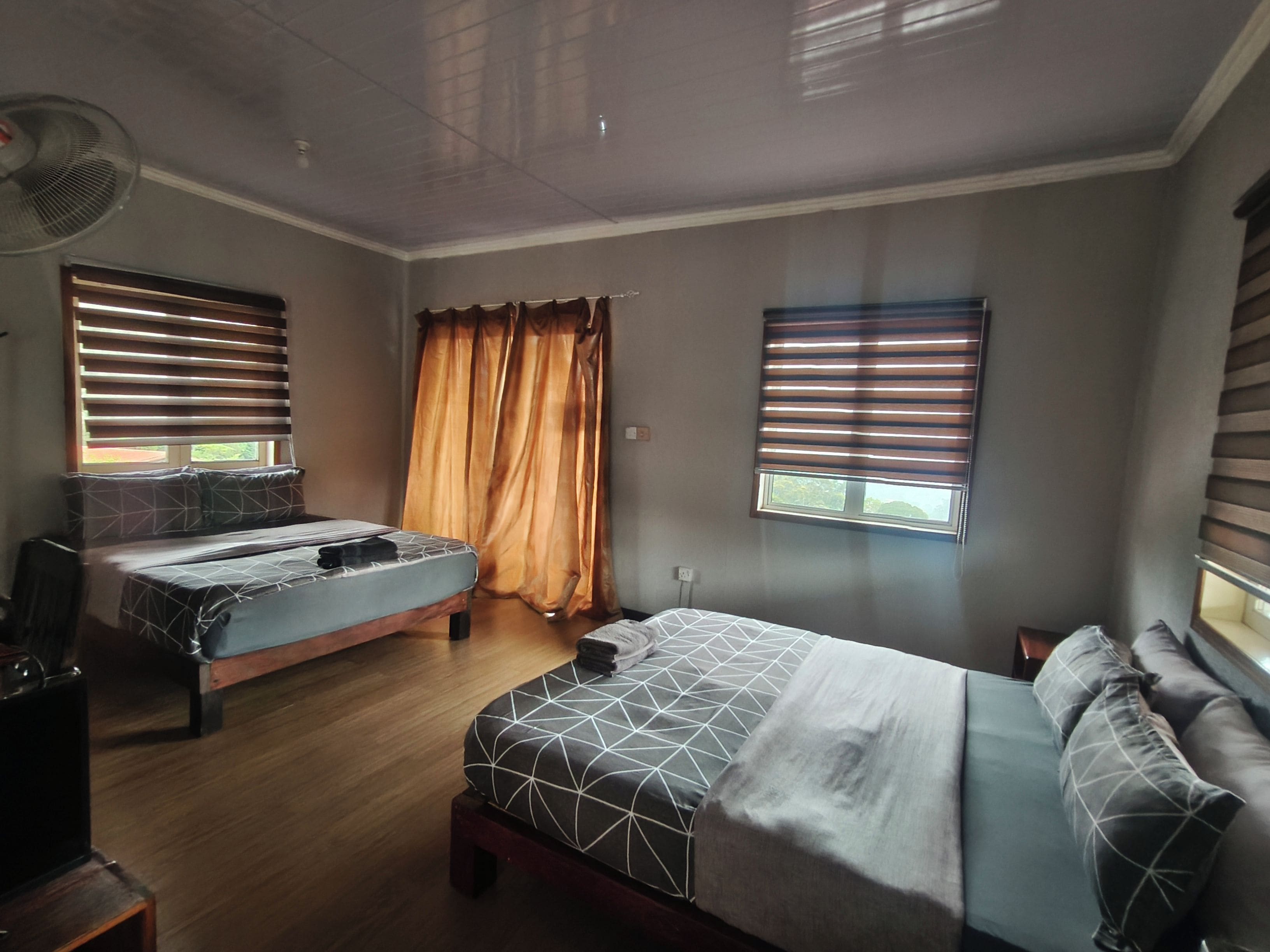 Een kamer met twee bedden, een bureau, een stoel, een ventilator en een raam met gordijnen.