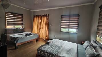 Een kamer met twee bedden, een bureau, een stoel, een ventilator en een raam met gordijnen.