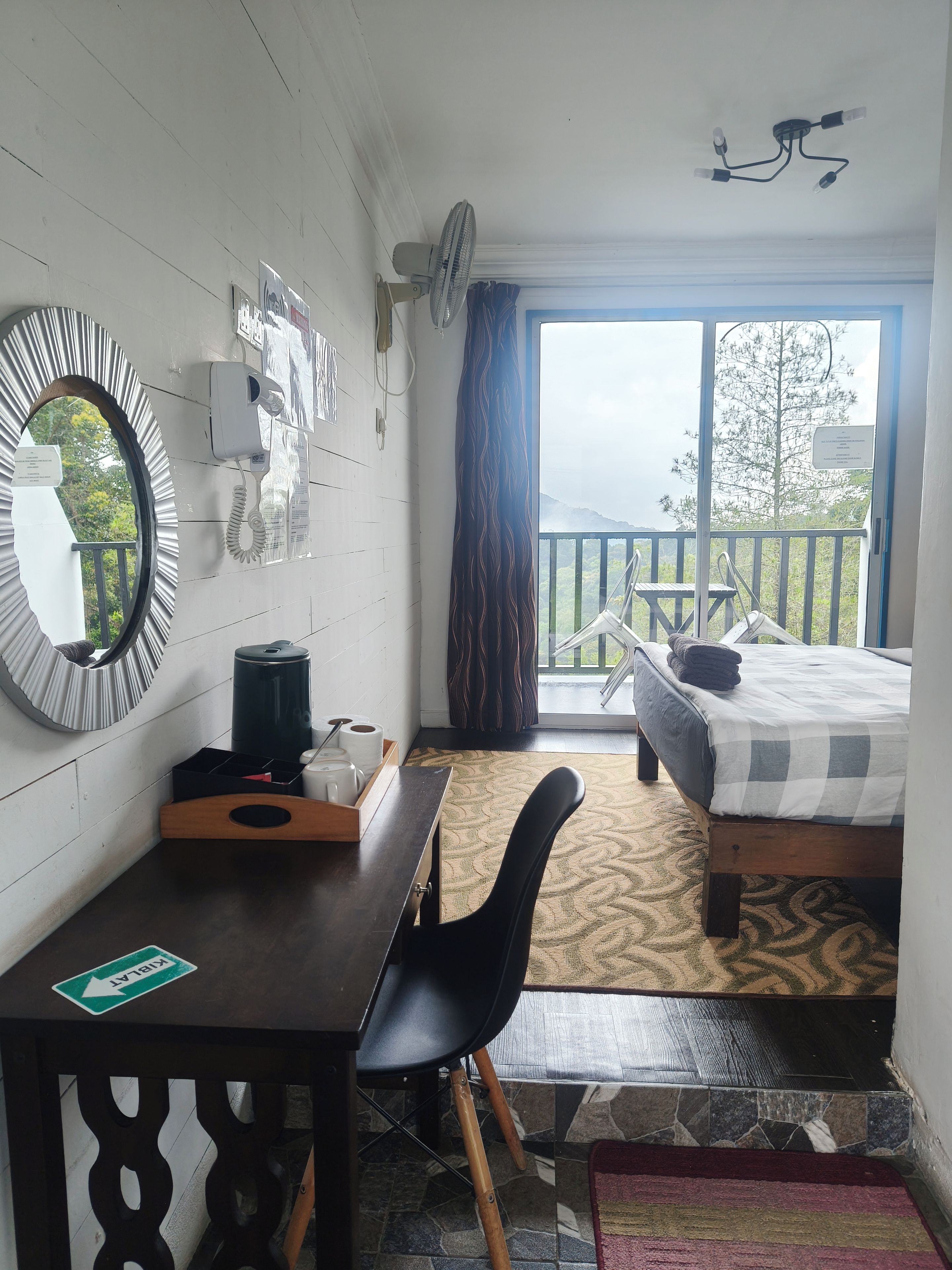 A room with a bed, a desk, a chair, a small table with a kettle and cups, a mirror, a telephone, a fan, and a balcony with a view of the mountains.