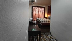 Una habitación de hotel pequeña con una cama, un escritorio, una silla, un ventilador y una cortina.