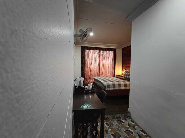 A compact hotel room with a bed, a desk, a chair, a fan, and a curtain. - Ceaser's Place (Ranau)