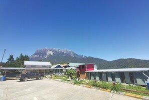 A parking area with a blue pickup truck, a building with a red section, and a mountain in the background. - Ceaser's Place (Ranau)