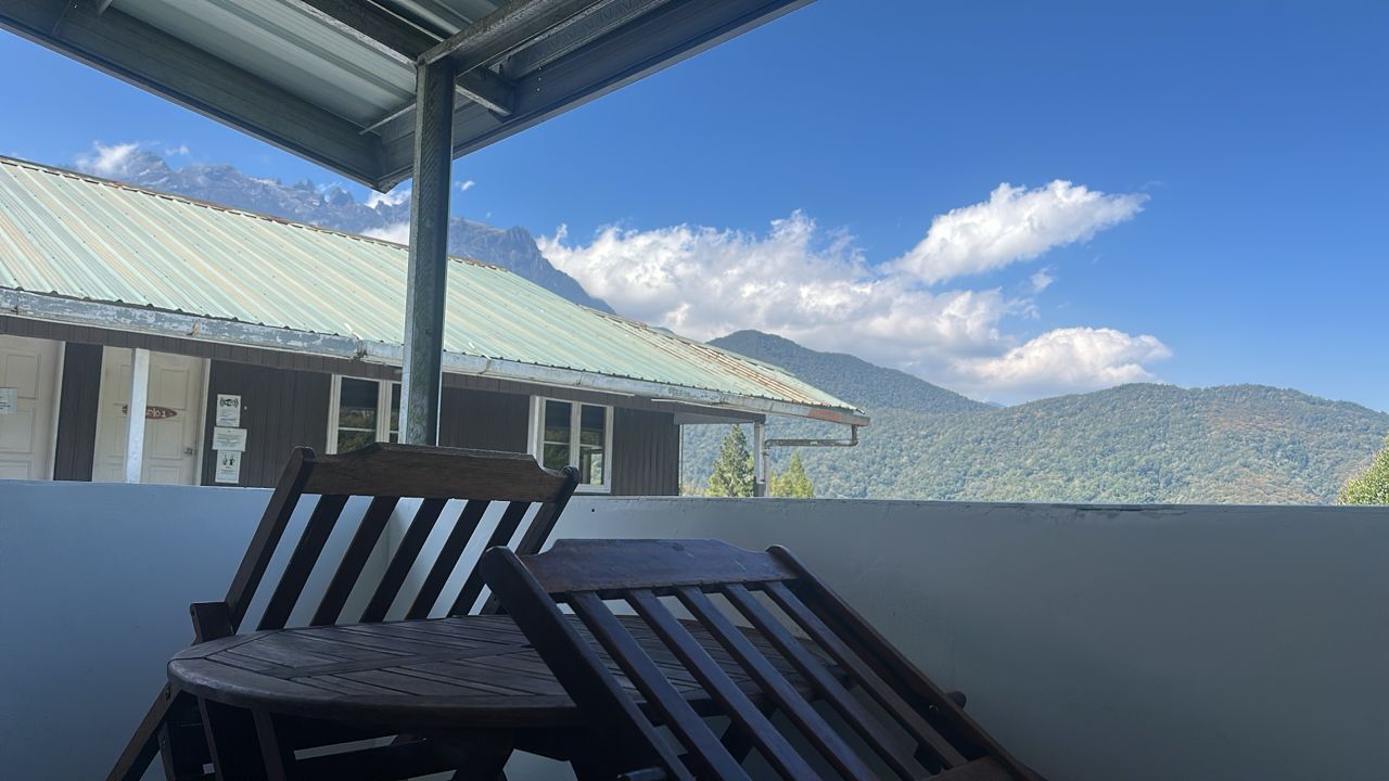Una silla y una mesa de madera en un balcón con vistas a montañas y un edificio con techo de metal corrugado.