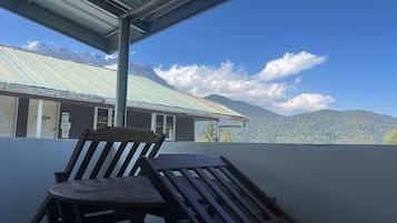 Una silla y una mesa de madera en un balcón con vistas a montañas y un edificio con techo de metal corrugado.