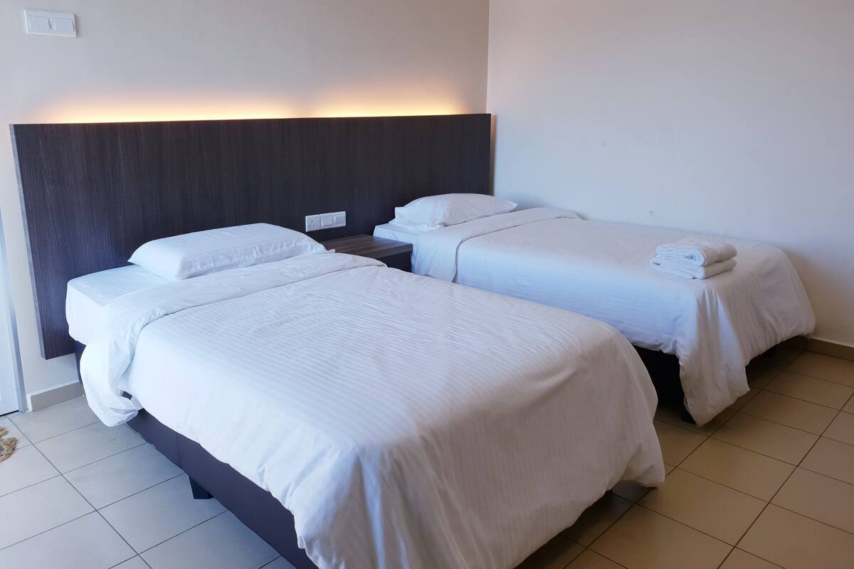 A hotel room with two single beds, a dark headboard, a nightstand, and a wall socket.