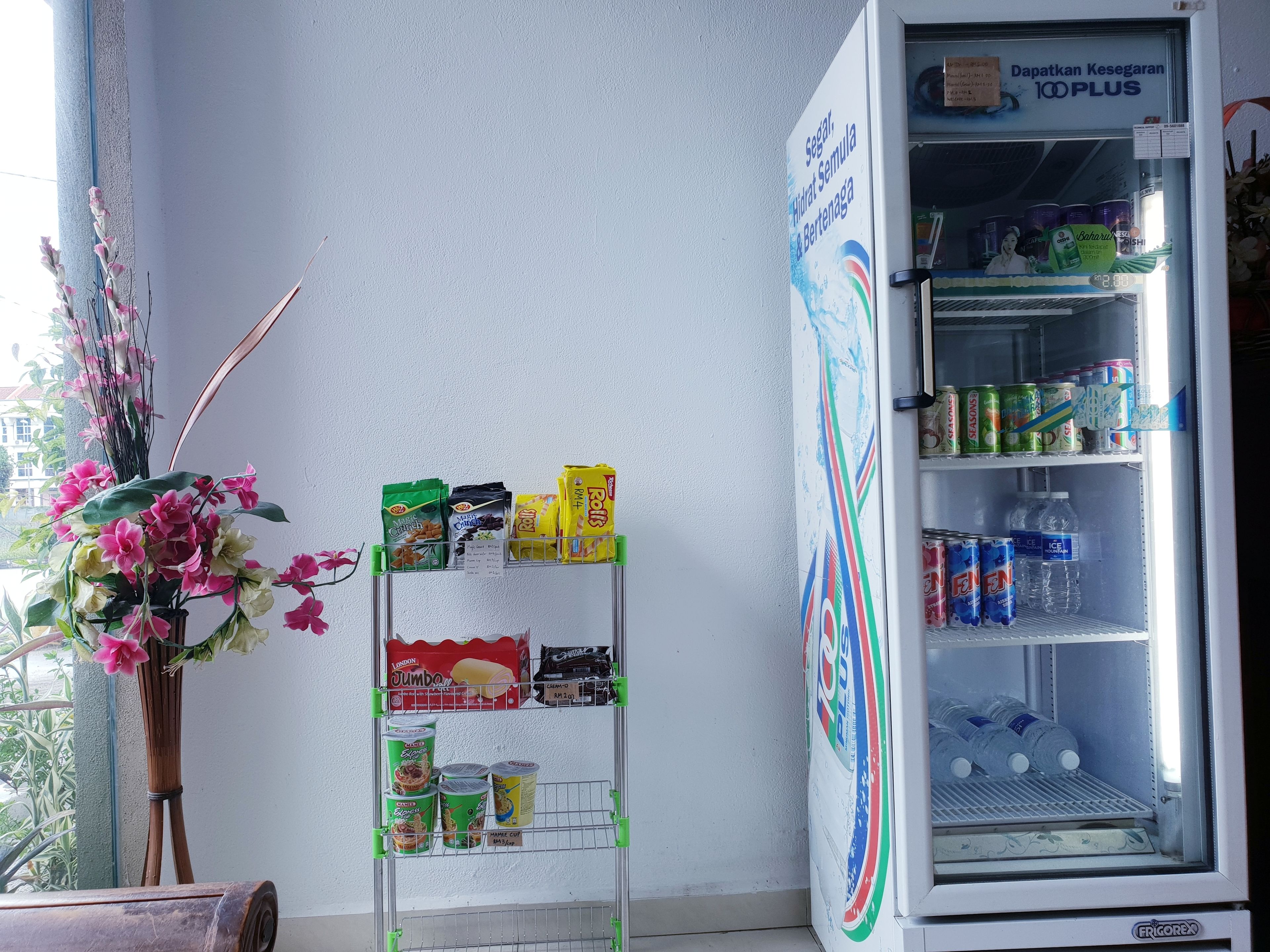 Una máquina expendedora llena de bocadillos y bebidas, un pequeño expositor con productos empaquetados, y un arreglo floral decorativo.