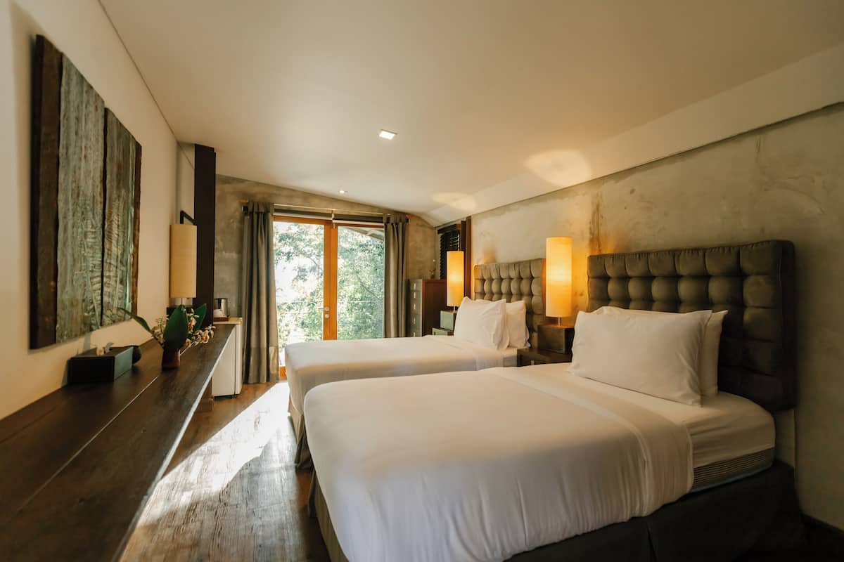 A hotel room with two beds, a large window with curtains, a wooden floor, and a wall-mounted artwork.