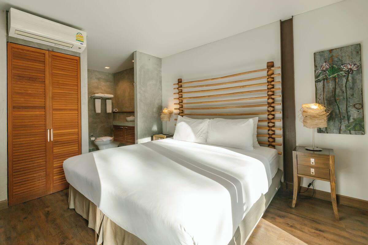 A bedroom with a wooden bed frame, a large white bedspread, a wooden nightstand with a lamp, and a wooden wardrobe.