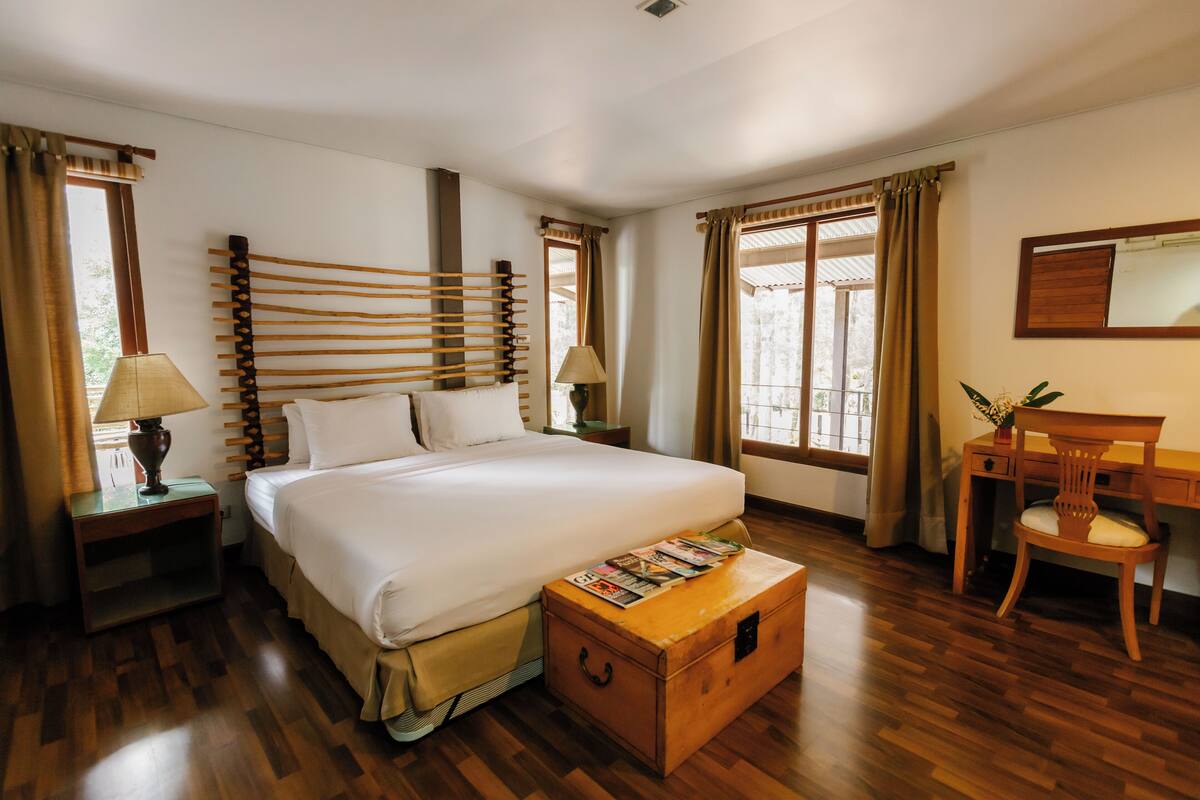 A hotel room with a large bed, wooden headboard, a desk with a chair, a wooden chest, and two nightstands with lamps.