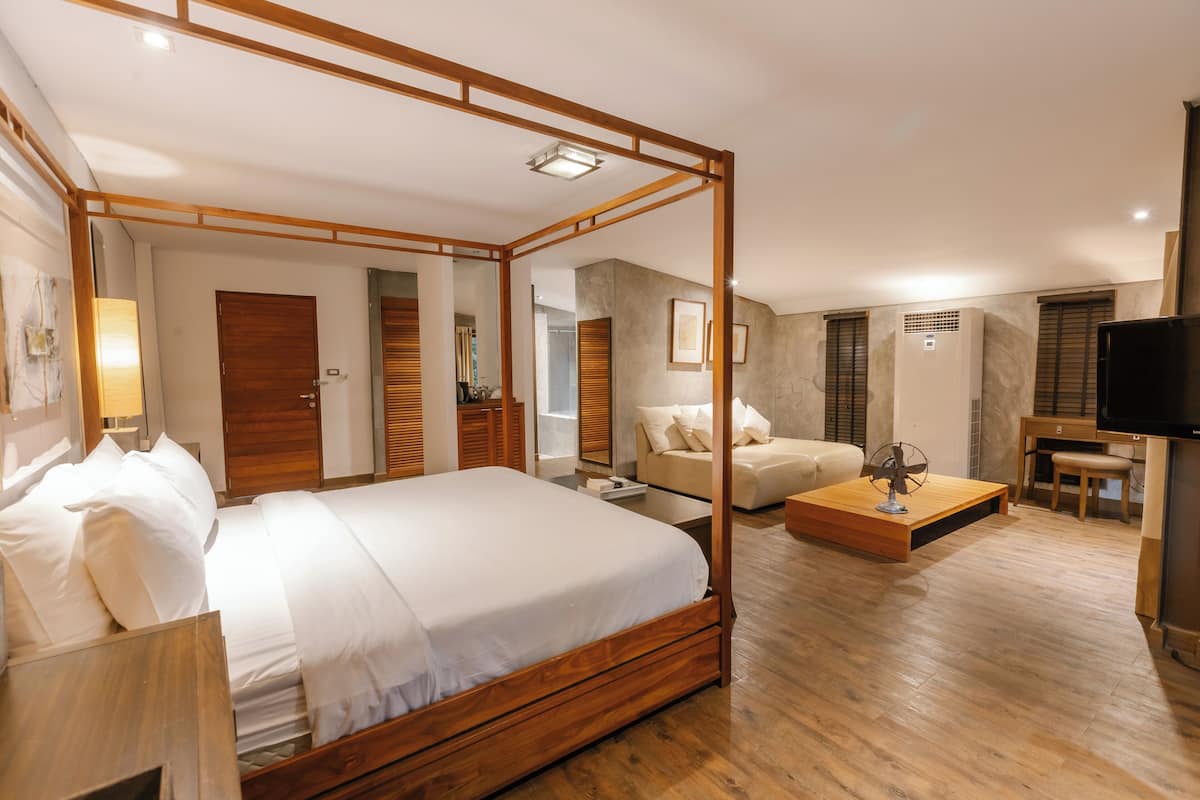 A four-poster bed with a wooden frame, a sofa, a TV, and a fan in a modern room with wooden flooring and a stone accent wall.