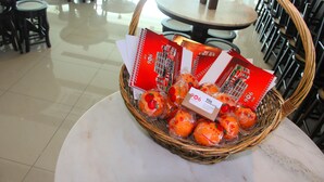 Un panier en osier contenant des oranges emballées et des cartes-cadeaux rouges, posé sur une table en marbre dans un coin repas agrémenté de plusieurs tables et chaises.