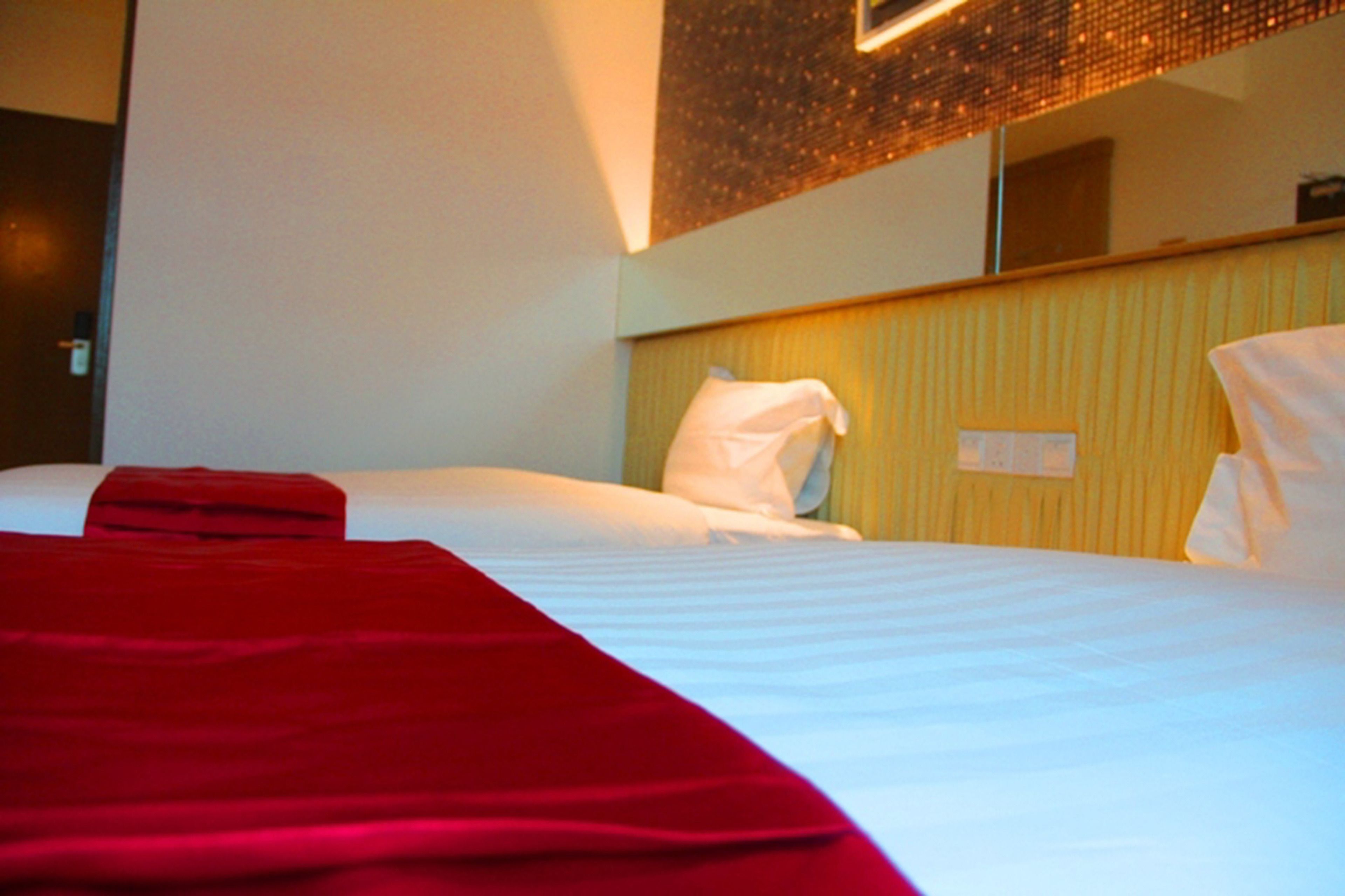 Une chambre d’hôtel avec un lit, une couverture rouge, un oreiller blanc et un miroir fixé au mur.