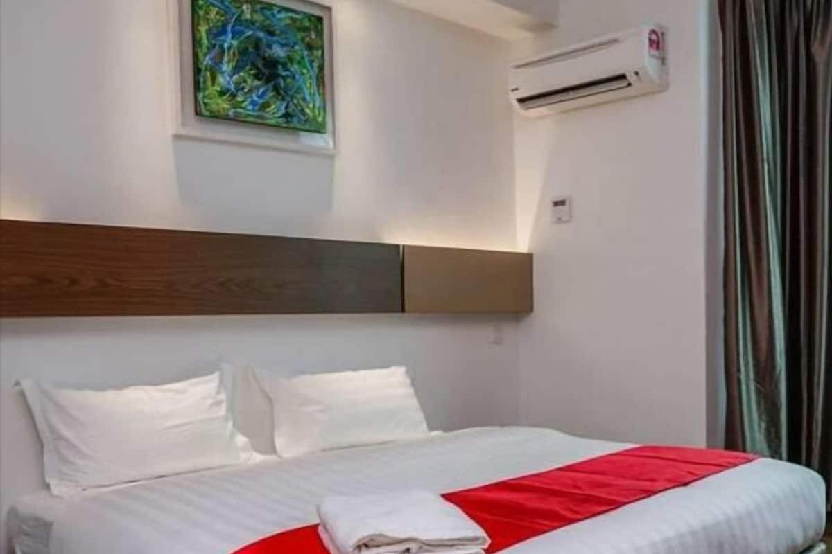 A hotel room with a large bed, white bedding, a red accent, an air conditioning unit, and a painting on the wall.