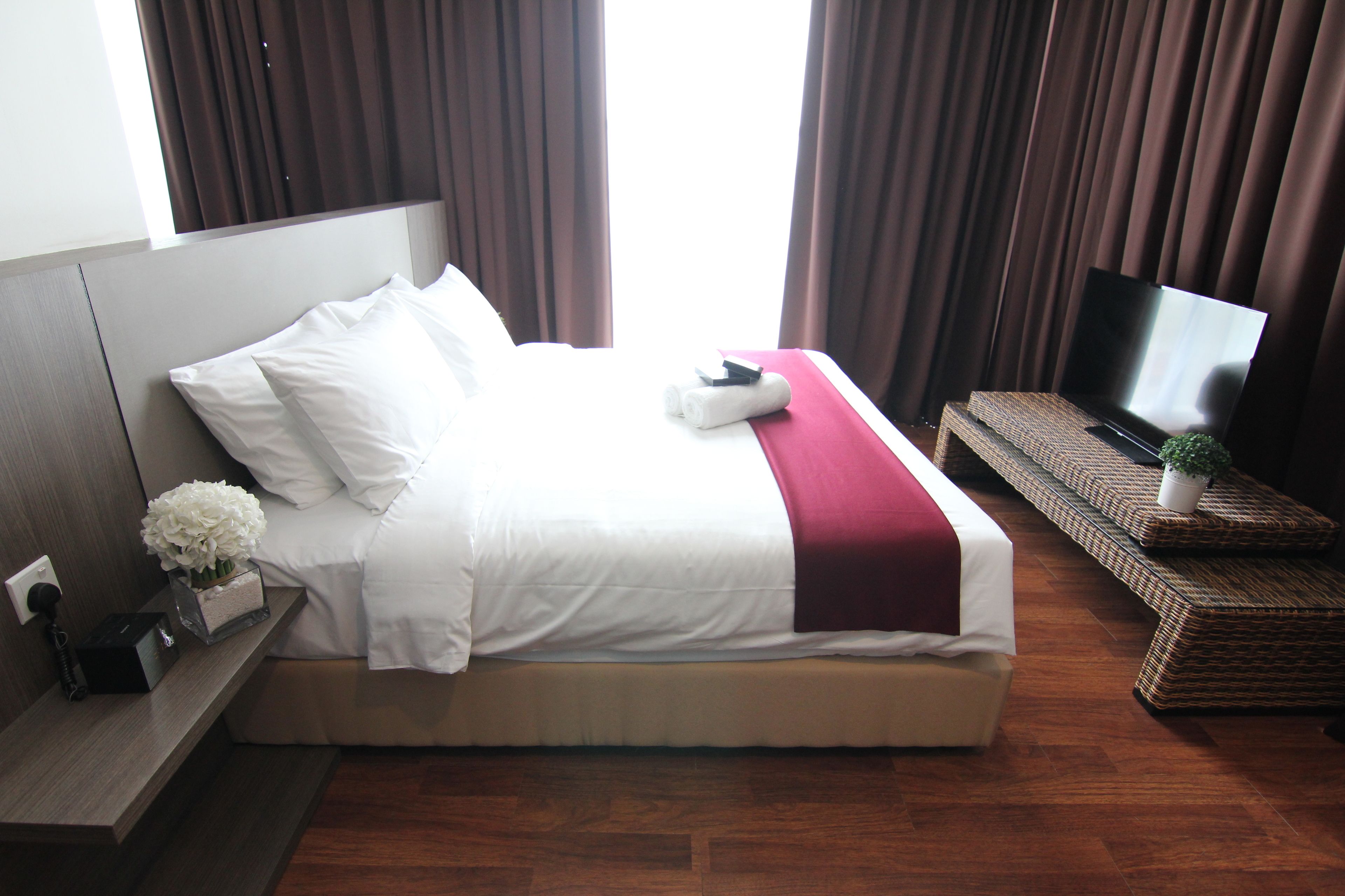 A neatly made bed with white linens and a red accent, a flat-screen TV on a stand, and a small potted plant on a wicker bench.
