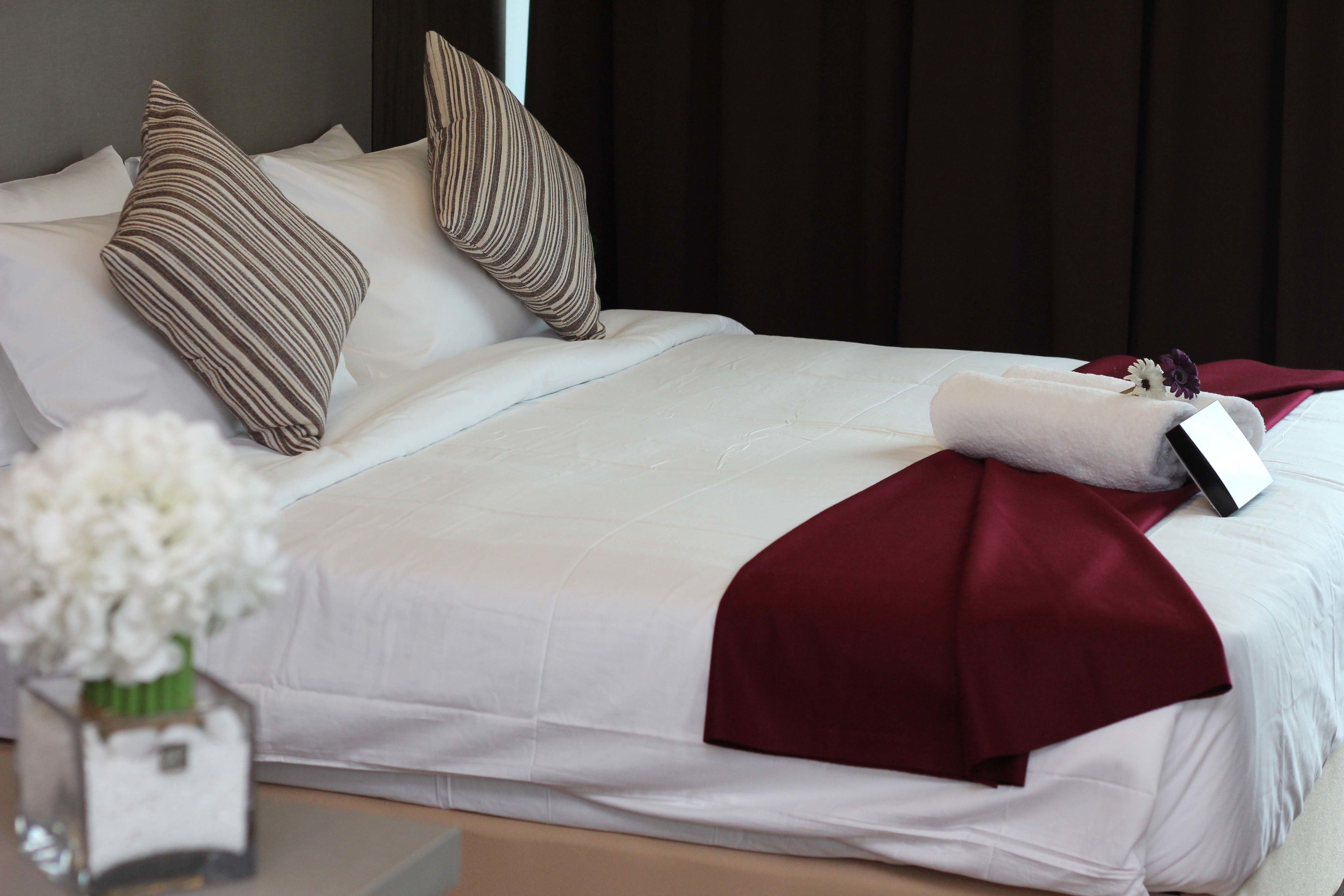 A neatly made bed with white and maroon bedding, striped pillows, and a small vase with white flowers on a bedside table.