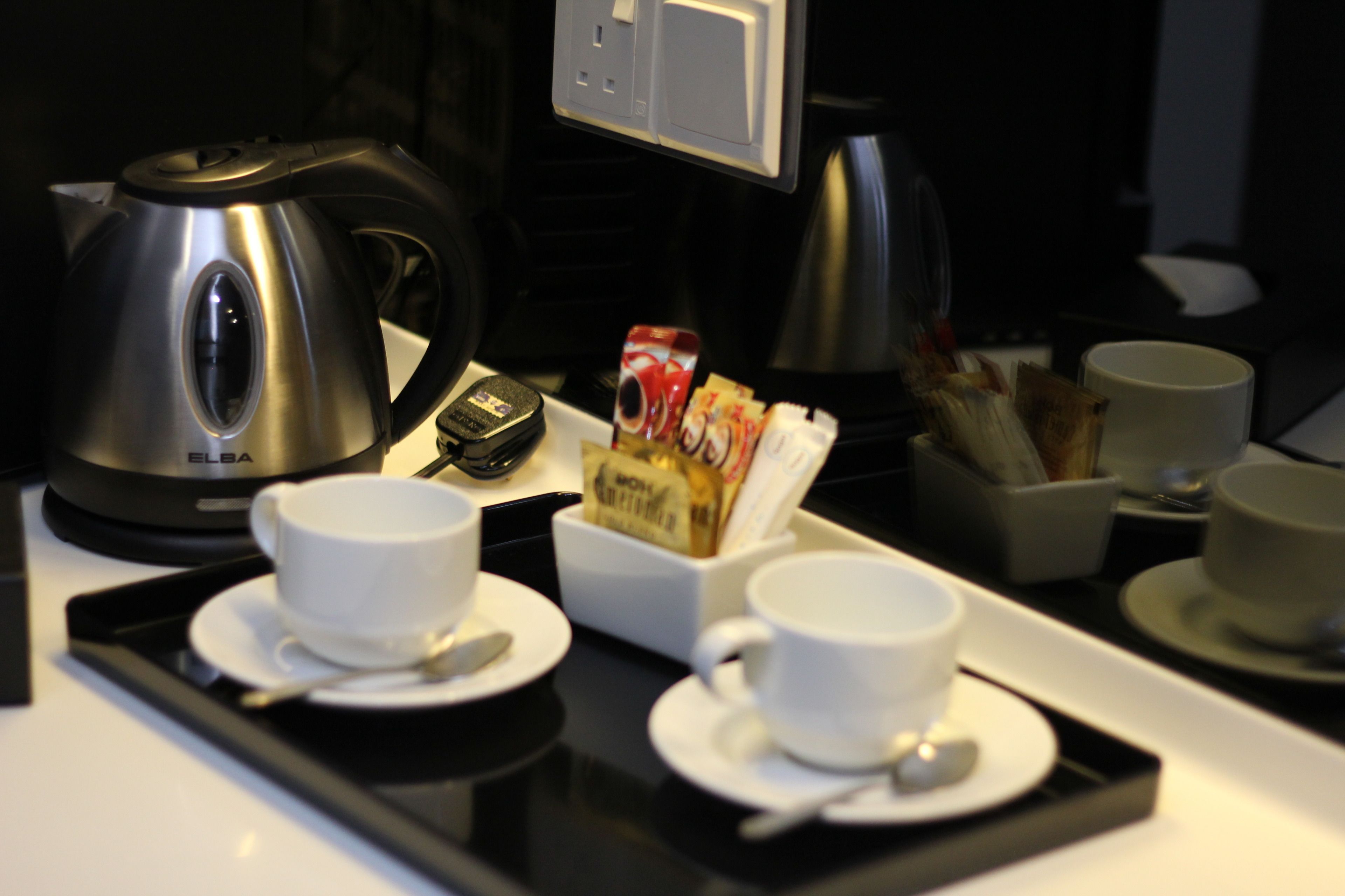 Preparación para el desayuno en una habitación de hotel, con una tetera, cafetera y una bandeja con tazas y una caja de cereales.