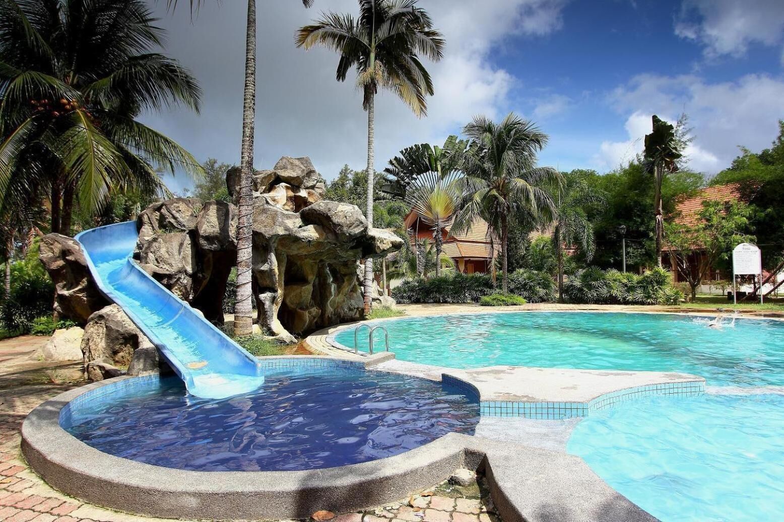 A swimming pool with a blue slide, surrounded by palm trees and a rocky area.