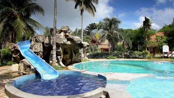 A swimming pool with a blue slide, surrounded by palm trees and a rocky area.