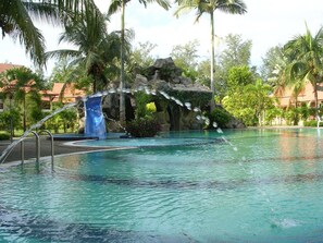 A swimming pool with a waterfall feature, surrounded by palm trees and a rocky backdrop.