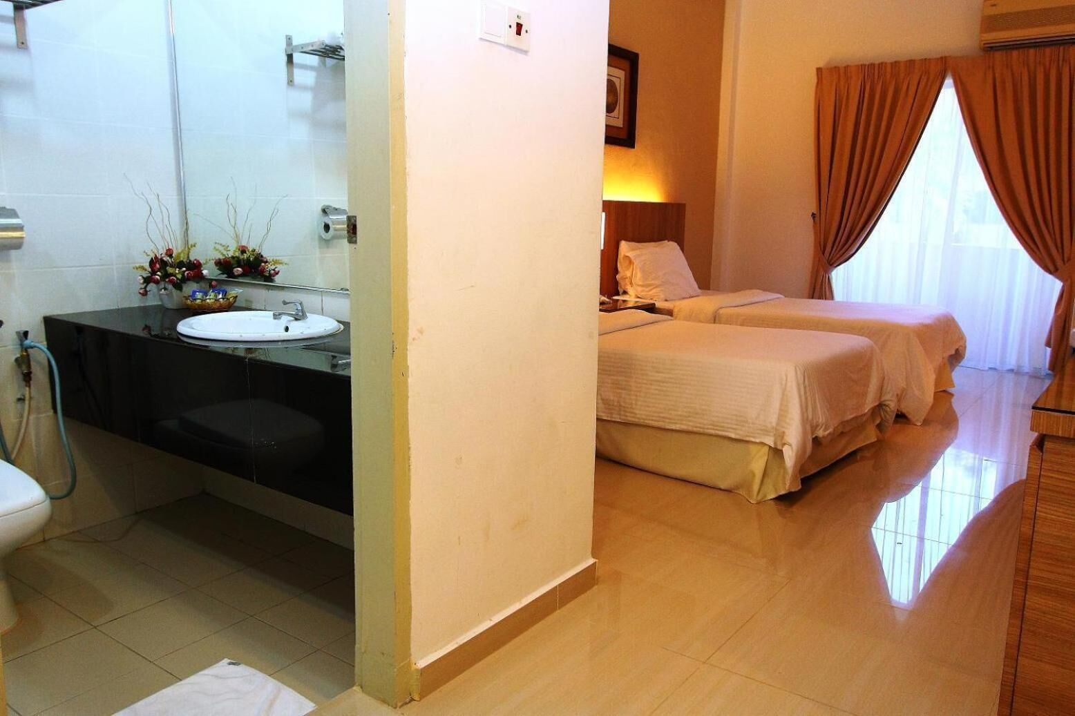 A hotel room with a bathroom featuring a sink, toilet, and shower. There are two beds with white bedding and a window with curtains.