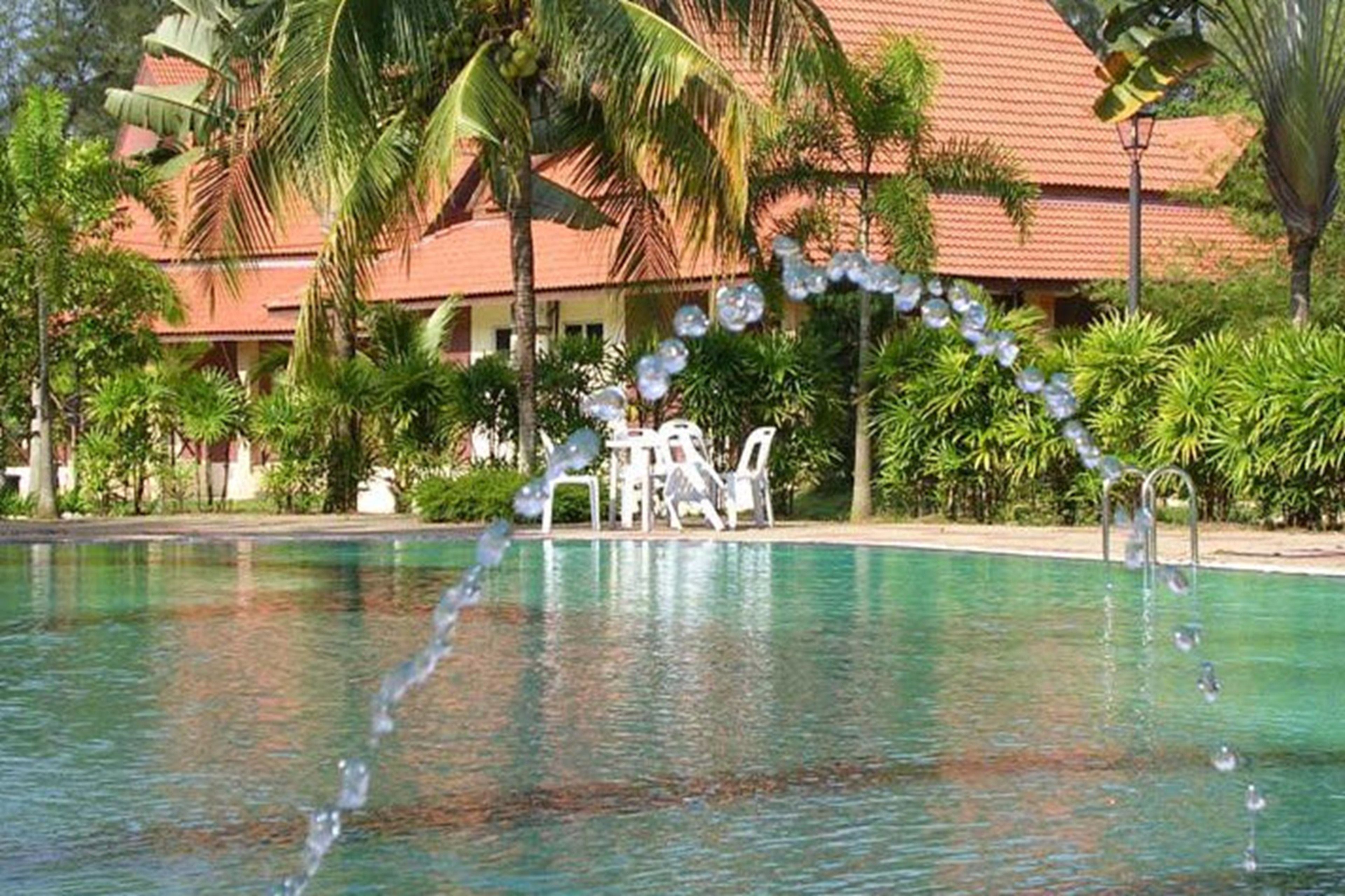 A swimming pool with a water feature, surrounded by palm trees and a building with a red roof.
