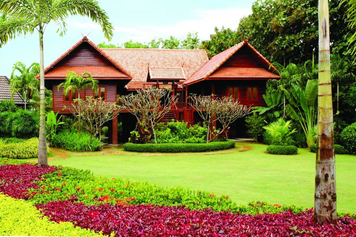 Una casa tradicional de madera con techo rojo, rodeada de exuberante follaje y un jardín cuidado con diversas plantas y flores.