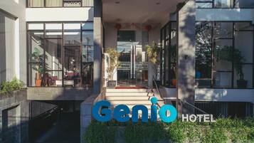 La entrada del Hotel Genio, con una vista despejada del interior gracias a amplios ventanales, una escalera que conduce a la entrada y plantas en macetas alrededor del área.