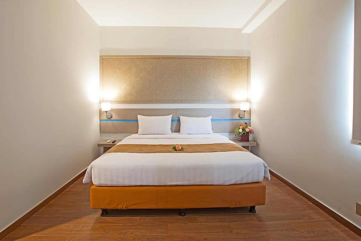 A hotel room with a large bed, wooden headboard, bedside lamps, and a small table with a vase of flowers.