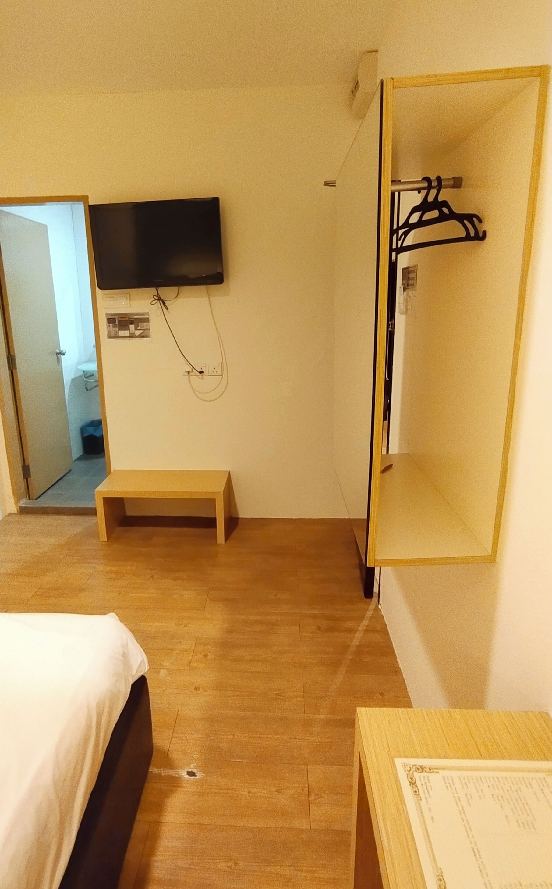 A hotel room with a bed, a wooden bench, a TV mounted on the wall, and a coat rack.