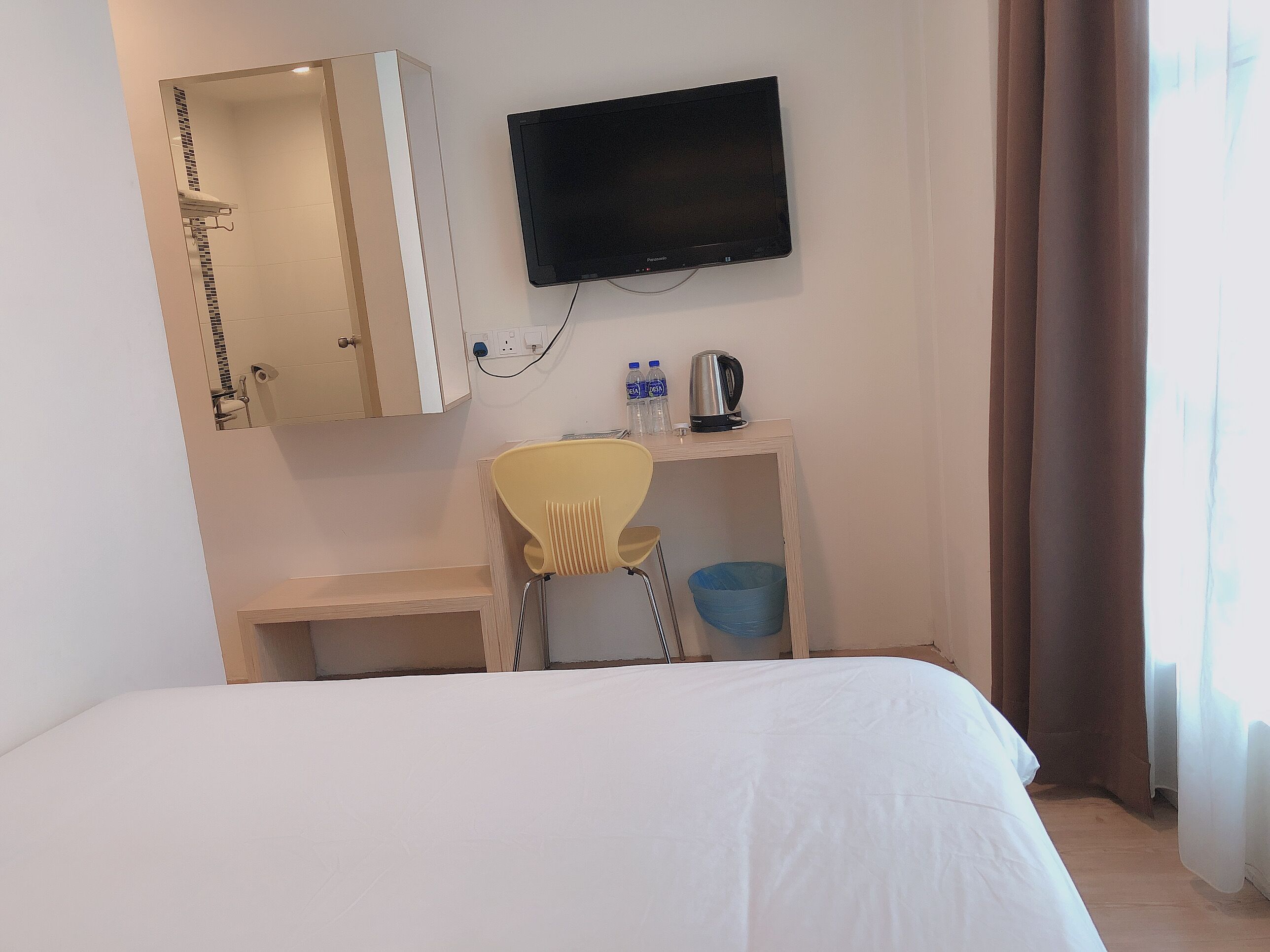 A hotel room with a bed, a desk with a chair, a television, a mirror, a sink, and a window with curtains.